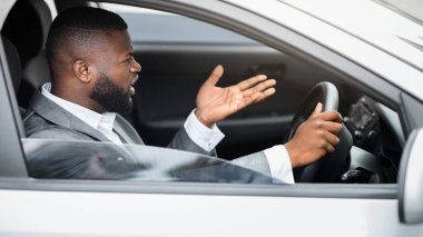 Öfkeli siyahi sürücü yolda kavga ediyor, trafiğe takılmış, yandan bakıyor. Stresli Afrikalı Amerikalı iş adamı araba sürerken bağırıyor ve el kol hareketi yapıyor, iş toplantısına gidiyor, boş yer var.