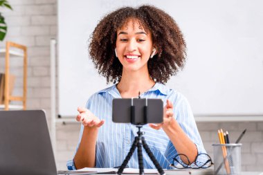 Evde yalnızlık çeken öğrenciler için çevrimiçi İngilizce dersi. Kablosuz kulaklıklı mutlu Afro-Amerikalı kadın akıllı telefonun web kamerasına bakıyor, Cvid-19 sırasında dizüstü bilgisayarla masada oturuyor.
