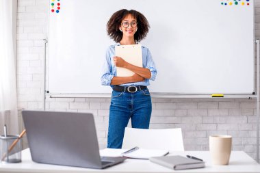 İngilizce öğretmeni ve ev dersleri online. Gözlüklü ve kablosuz kulaklıklı gülümseyen Afrikalı Amerikalı kadın oturma odasında dizüstü bilgisayarı ve not defteri olan beyaz tahta ve masanın yanında duruyor.