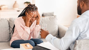 Psikolog ofisteki zihinsel terapi seansı sırasında depresyondaki siyahi kadını rahatlatıyor, bir bardak su sunuyor, profesyonel yardıma ihtiyacı olan koltukta oturan Afrikalı kadın.