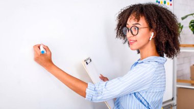 Online eğitim öğretmeni. Çekici Afro-Amerikan kadın gözlüklü ve kablosuz kulaklıklı İngilizce kurallar yazıyor ve salgın sırasında öğrencilere video yazıyor.