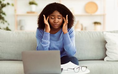 Aşırı çalışma ve teslim tarihi kavramı. Dizüstü bilgisayarda çalışan, kanepeye oturan, şakaklara dokunan ve masaj yapan stresli Afro-Amerikan kadın. Siyah kadın baş ağrısı çekiyor, stresi azaltıyor, düşünüyor.