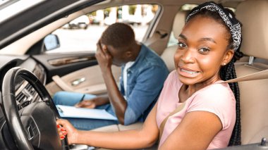 Eğitmenle okul arabası süren genç siyahi bayan, hata yapıyor ya da sınavdan kalıyor, çizelgeyle oturup kafasına dokunuyor, yandan bakıyor, boşluğu kopyalıyor.