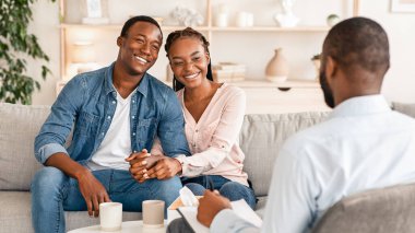 Etkili Evlilik Terapisi. Mutlu Afrikalı çift danışman ofisindeki koltukta oturuyor, el ele tutuşuyorlar ve gülümsüyorlar, terapistin dikiz açısı, boş alan.