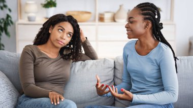 Dishonest african american woman listening to her happy girlfriend sharing feelings, home interior, copy space. Envious black lady pretending been a good friend, sitting on sofa in living room
