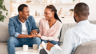 Uzlaşma konsepti. Evlilik terapisti ofisinde el ele tutuşan mutlu siyah çift, tartışmadan sonra tekrar bir araya geliyor, birbirlerine gülümseyerek koltuğa oturuyor, seçici bir şekilde odaklanıyorlar.