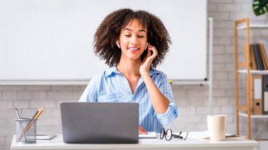 Uzaktan İngilizce öğretmeni. Çekici Afrikalı Amerikalı kablosuz kulaklıklı genç bir kadın dizüstü bilgisayara bakıyor ve içerideki öğrencilerle iletişim kuruyor.