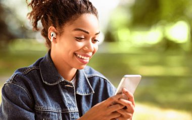 Neşeli Siyah Kadın Cep Telefonu ve Kulaklık Kullanıyor Dışarıda Park 'ta Oturan Kulaklıklar. Müzik Uygulaması Konsepti