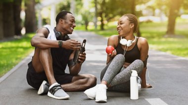 Neşeli Afrikalı Amerikalı çift dışarıda egzersiz yaptıktan sonra dinleniyor, şehir parkında yolda oturuyor, su içiyor ve sağlıklı atıştırmalıklar yiyorlar.