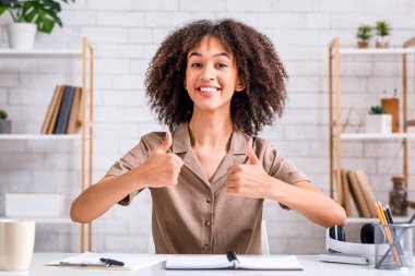 Modern ev blogu yazarı, harika eleştiri ve hobi. Gülümseyen Afrikalı Amerikalı kadın başparmağını kaldırıyor ve kameraya bakıyor, minimalist oturma odasında dizüstü bilgisayarla masada oturuyor, boş yer var.