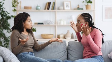 Emotional african american ladies best friends fighting while sitting on couch at home, copy space between them. Angry black women screaming and gesturing, having crisis in friendship
