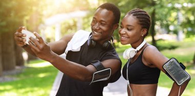 Siyah milenyum çifti, dışarıda spor yaparken akıllı telefondan selfie çekiyor, kameraya bakıyor ve gülümsüyor.