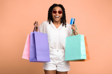 Kredi kartıyla alışveriş yapan Afrikalı Kız ve Satış konusunda heyecanla gülümseyen alışveriş torbaları, Bej Stüdyo arka planı. Alışveriş koliği.