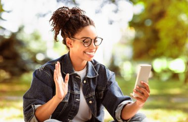 Akıllı telefon kullanan mutlu Afrikalı Amerikalı kadın dışarıda park yerinde oturmuş cep telefonuna el sallıyor. Uzak Online İletişim Konsepti. Seçici Odaklanma
