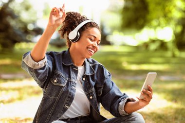 Kulaklıklı neşeli siyahi bayan dışarıda parkta oturmuş online müzik dinliyor. Afrikalı Öğrenci Sevdiği Şarkılar, Sonbahar Çalma Listesi ve Haftasonu Gevşeme Konsepti