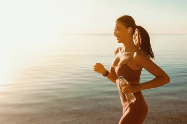 Spor hayattır. Yanında kulaklık takan bir şişe maden suyu ile deniz kenarında koşan kadın koşucunun portresi.