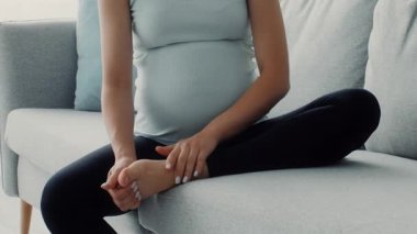Evdeki kanepede rahat bir şekilde oturan genç bir kadın şişmiş ayaklarına nazikçe masaj yapıyor. Rahatlamış görünüyor ve rahat ortamında sakin bir anın tadını çıkarıyor..
