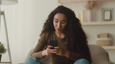 Sıcak bir yuva ortamında akıllı telefonuyla meşgul olan kıvırcık saçlı genç bir Arap kadın gülümsüyor. Atmosfer gündüzleri rahat bir yaşam tarzını yansıtıyor..