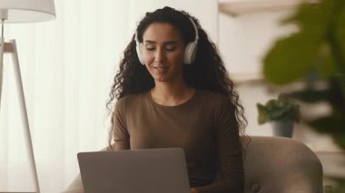 Genç bir Orta Doğulu kadın evde, kulaklık takıyor ve laptopuyla meşgul. Rahat bir ortamda, teknolojiyi kucaklayan sıradan bir yaşam tarzı sunuyor..