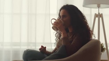 Genç bir Orta Doğulu kadın oturma odasında rahatça oturup sıcak bir şeyler içiyor. Rahatlamış ve meşgul görünüyor, boş zamanlarında evde huzurlu bir yaşam tarzını somutlaştırıyor..