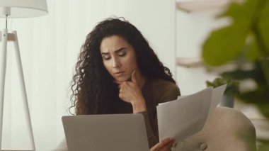 Kıvırcık saçlı genç bir Orta Doğulu kadın kanepeye rahatça oturur ve elinde bazı kağıtlar tutarken dikkatle dizüstü bilgisayarına odaklanır. Evdeki göreviyle meşgul görünüyor..