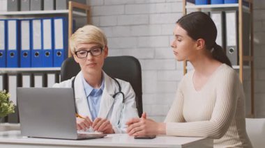 Beyaz önlüklü bir beslenme uzmanı kişisel bir muayene sırasında bir kadın hastayla beslenme seçeneklerini tartışıyor. Modern klinikte ahşap raflar ve bir dizüstü bilgisayar bulunuyor.