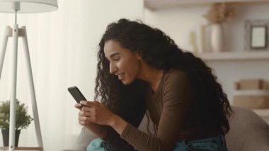 Genç bir Ortadoğulu kadın evdeki rahat bir kanepede dinleniyor, akıllı telefonuyla konuşurken gülümsüyor. Gündelik tarzı, boş zamanlara ve bağlantılara odaklanmış çağdaş bir yaşam tarzını yansıtıyor..