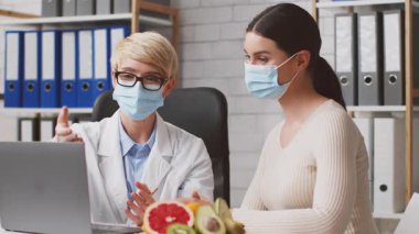 Beyaz önlüklü bir diyetçi klinik ortamındaki bir müşteriyle beslenme planlarını tartışıyor. Her ikisi de maske takıp dizüstü bilgisayara odaklanırken sağlıklı yiyecekler masada görülebiliyor..