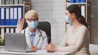 Bir beslenme uzmanı, zeki ve düzenli bir klinikte kadın bir müşteriyle görüşüyor. Maskeli sağlık protokollerini korurken beslenme seçeneklerini tartışıyor ve referans için bir dizüstü bilgisayar açık tutuyorlar..