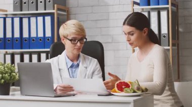 Klinikte diyetçi bir kadın hasta ile kişisel beslenme planını tartışıyor. Taze meyveler, sağlıklı besin seçeneklerini ve beslenme rehberliğini vurgulamak üzere masada mevcuttur..