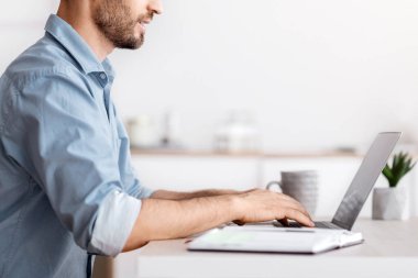 Beyaz ofisteki işyerinde oturan yakışıklı bir iş adamı, dizüstü bilgisayarda çalışıyor, klavyeyle yazı yazıyor, e-postaları cevaplıyor, müşterilerle mektuplaşıyor, yan görünümü kopyalama alanı ile