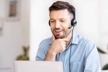 İş Eğitimi. Kulaklıklı yakışıklı genç işadamı modern ofiste dizüstü bilgisayarda webinar izliyor, çevrimiçi eğitim alıyor, bilgisayar ekranına ilgiyle bakıyor, masada oturuyor, boşluğu kopyalıyor.