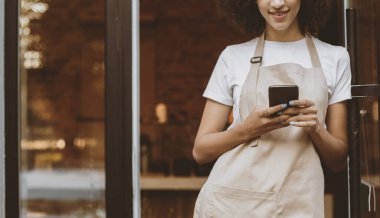 Modern servis, kafeteryadan içki siparişi ve teslimatı için çevrimiçi uygulama. Önlüklü mutlu Afrikalı Amerikalı bayan sabahları kafenin ön kapısının yanındaki akıllı telefona bakar.