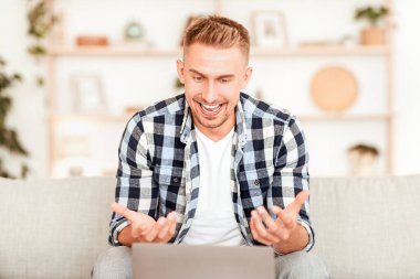 Çevrimiçi İletişim Konsepti. Bilgisayarda sanal görüntülü konuşma yapan genç bir adamın portresi. Konuşuyor, webcam 'e bakıyor ve el kol hareketi yapıyor. Adam bilgisayarla sohbet ediyor, evdeki koltukta oturuyor.