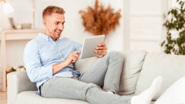 Teknoloji Konsepti. Modern dairesindeki koltukta oturan dijital tablet kullanan gülümseyen adamın portresi, fotokopi alanı. Mutlu adam haberleri okuyor, internette geziniyor, oturma odasındaki kanepede dinleniyor.