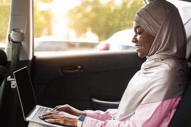 Çevrimiçi İş. Gülümseyen Müslüman iş kadını arabanın arka koltuğunda dizüstü bilgisayar kullanıyor, internette çalışıyor, tesettüre giriyor, İslami Kadın Girişimci Kişisel Sürücüyle Ofise Gidiyor