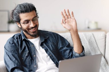 Neşeli genç bir Arap 'ın evde dizüstü bilgisayarında görüntülü araması kameraya el sallaması oturma odasında dinlenirken ailesiyle konuşması internet iletişiminin tadını çıkarması, boşluğu kopyalaması.