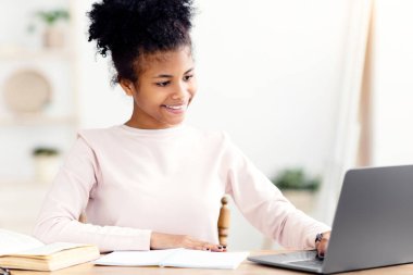 Afrikalı genç kız dizüstü bilgisayarda ödev yapıyor. İnternette çalışıyor. Evde masa başında oturuyor. Uzaktan Öğrenme Konsepti