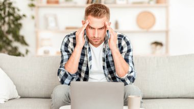 Aşırı çalışma ve teslim tarihi kavramı. Stresli sıradan bir adam dizüstü bilgisayara bakıyor kanepeye oturuyor, şakaklara dokunuyor.