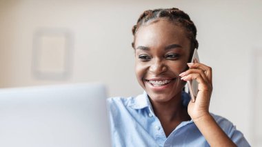 İş İletişimi. Neşeli Siyah Kadın Girişimci Cep telefonuyla konuşuyor ve iş yerinde dizüstü bilgisayar kullanıyor.