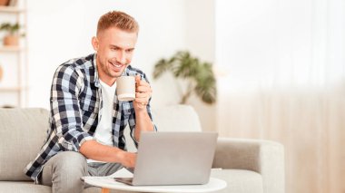 Uzaktan Çalışma ve Çalışma Konsepti. Oturma odasında dizüstü bilgisayarda yazan, kanepede oturan, kahve içen ve evdeki kişisel bilgisayarı kullanan gülümseyen adamın portresi.