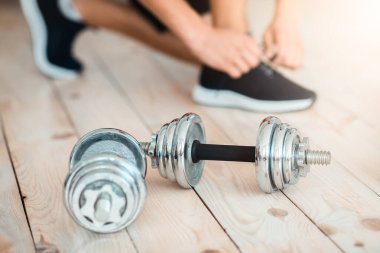 Evde dambıllarla egzersiz yapmak. Ayakkabı bağlayan adam, yerde protein içeceği ve spor malzemeleri için bardağa yakın, kırpılmış, kapat.