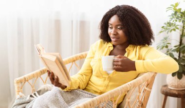 Hafta sonu eğlencesi. Gülümseyen siyah bir kadının portresi kitap okuyor, sıcak kahve içiyor, elinde fincanla hasır bir sandalyede battaniyeyle oturuyor. Uzayı kopyala, arkaplanı bulanıklaştır. Dinlenme ve Rahatlama Kavramı