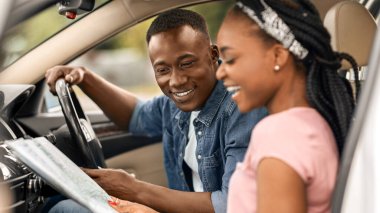 Araba yolculuğu, otomobil konseptiyle seyahat. Mutlu milenyum siyahi kadını, erkek arkadaşına kağıt harita üzerinde seyahat edeceği yeri gösteriyor, otomobilde ön koltuklarda oturuyor, gelecekteki yolculuğunu, kenar manzarasını tartışıyor.