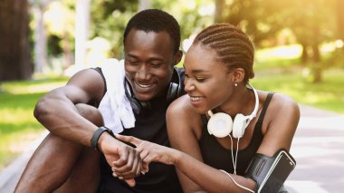 Neşeli siyah çift parkta koştuktan sonra dinleniyor, patikada otururken akıllı spor takip cihazlarını kontrol ediyor, dışarıda antrenmanların keyfini çıkarıyor, boş alan arıyor.