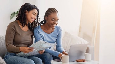 Two cheerful black women checking hotels online, using laptop, looking at map, home interior, free space. Excited african american ladies travelers getting ready for vacation, buying travelling tour