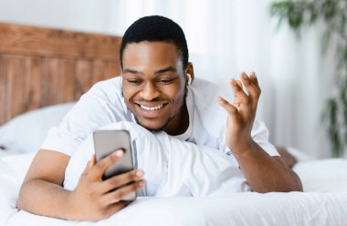 Uzaktan iletişim için akıllı telefon ve kulaklık kullanarak hafta sonları evdeki yatak odasında uzanıp kulaklık takan mutlu siyahi adam. Seçici Odaklanma
