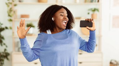 Müzik aşığı. Heyecanlı Afrikalı Amerikalı kadın portresi şarkı söyleyen ve cep telefonunu mikrofon olarak tutan, dans eden ve kablosuz kulaklık takan. Duygusal siyahi kadın aletinde karaoke uygulaması kullanıyor.