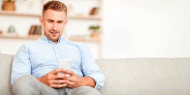 Boş zaman kavramı. Akıllı telefonunu kullanan yakışıklı bir adamın portresi, evdeki koltukta oturan bulanık arka plan, kopyalama alanı. İnternette sohbet eden, sosyal medyada gezinen, internet mesajları atan bir adam.