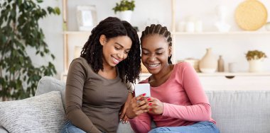 Joyful black girlfriend sitting on sofa in living room and using smartphone, copy space. Two happy african american young women reading beauty blog together, spending weekend at home